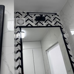 Oleh Tanchak Top Remont - Nowoczesna łazienka z lustrem w czarnej ramie, otoczonymi płytkami w zygzak. Widoczny wentylator i oświetlenie nad lustrem. Białe marmurowe płytki na ścianach.