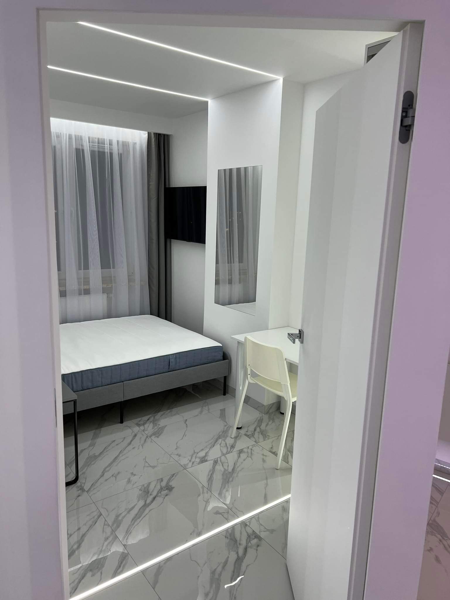 Nowoczesna sypialnia z białymi ścianami, marmurową podłogą, łóżkiem z szarą ramą, biurkiem i oknem z zasłonami, widziana zza uchylonych białych drzwi.