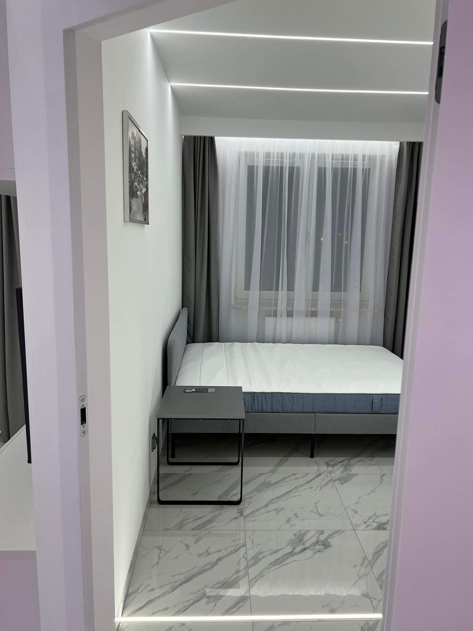 Sypialnia z łóżkiem kontynentalnym, marmurową podłogą i nastrojowym oświetleniem LED wzdłuż sufitu i listew przypodłogowych, widok zza framugi drzwi.