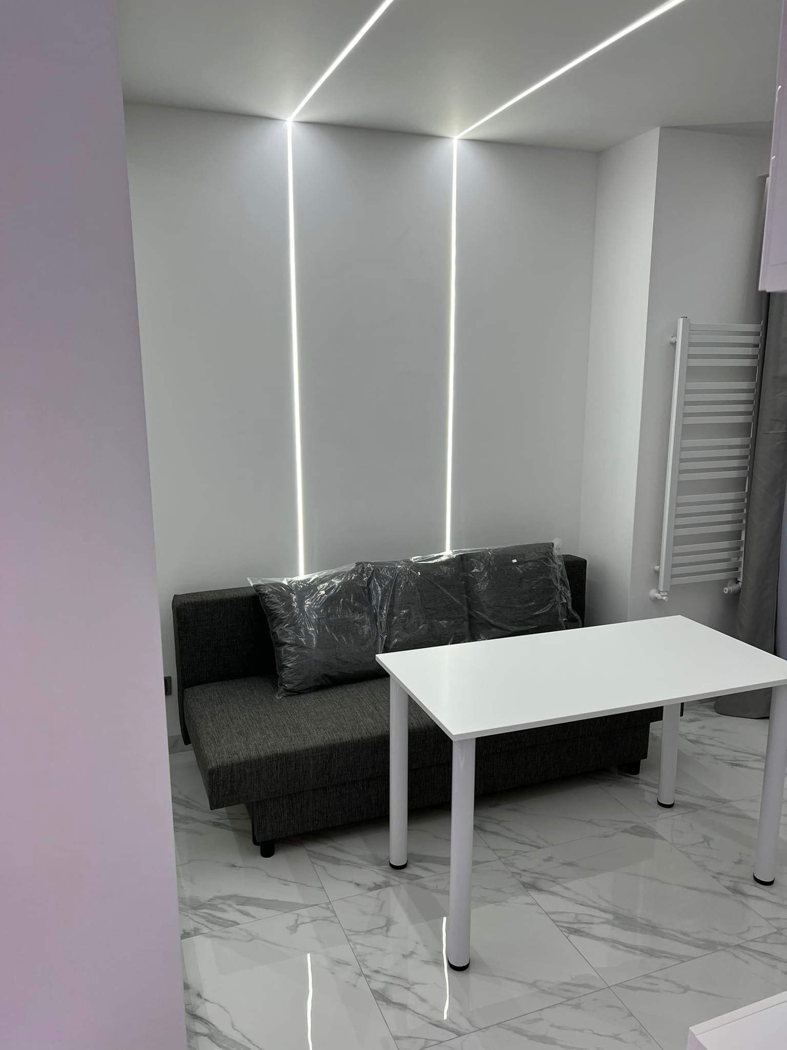 Nowoczesny salon z szarą rozkładaną sofą z poduszkami w folii, białym stołem i pionowymi liniami LED na ścianie, uzupełniony grzejnikiem drabinkowym.