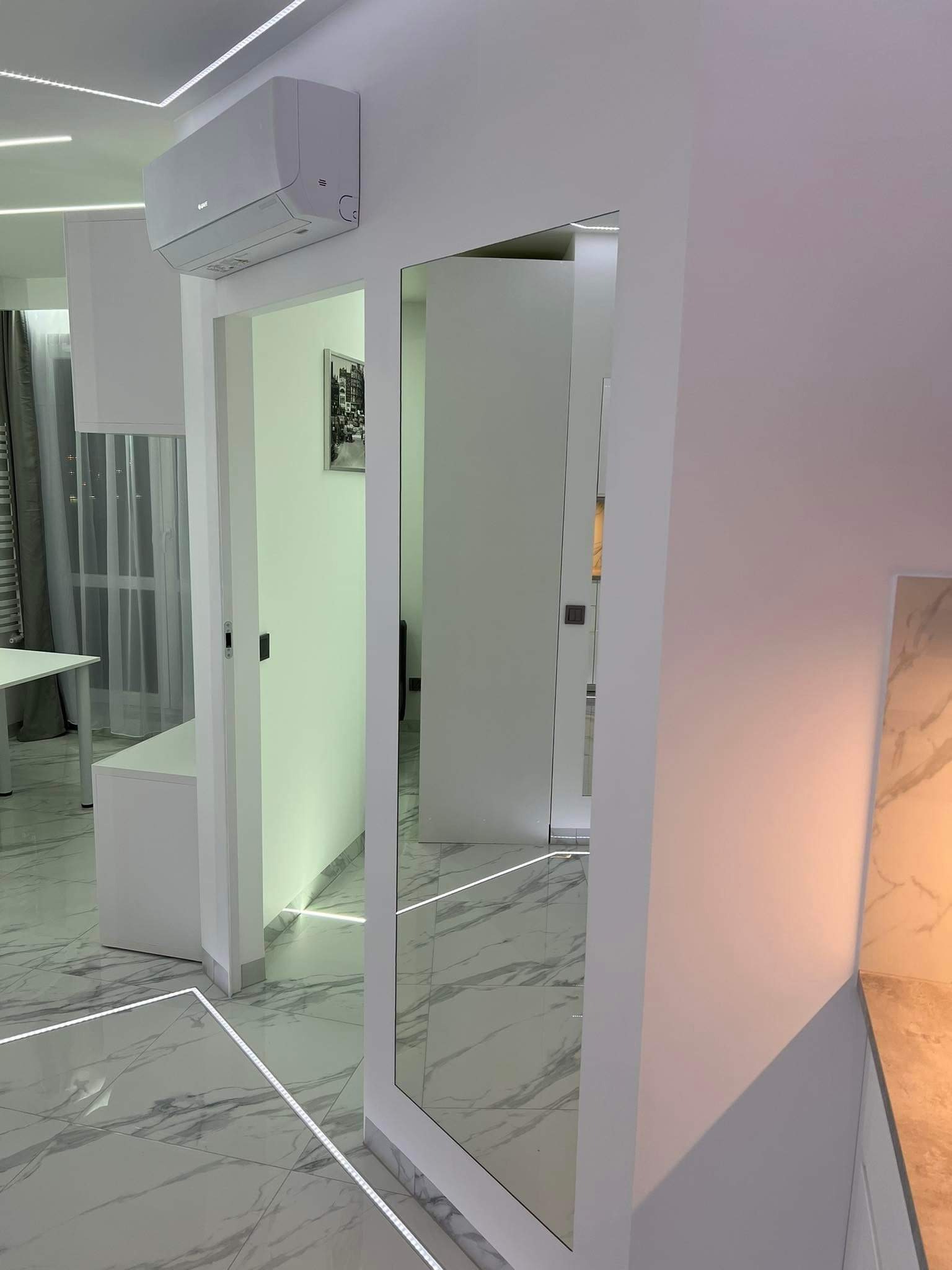 Nowoczesne wnętrze: białe ściany, marmurowe płytki podłogowe z podświetleniem LED, lustro w białej ramie i klimatyzator na ścianie.