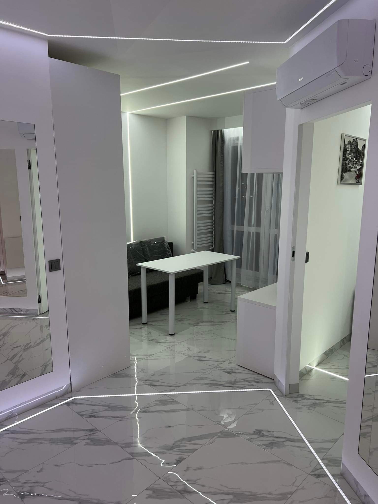 Nowoczesne wnętrze mieszkania z białymi ścianami, marmurową podłogą, oświetleniem LED wzdłuż krawędzi, klimatyzacją i widokiem na pokój z szarą sofą i białym stołem.