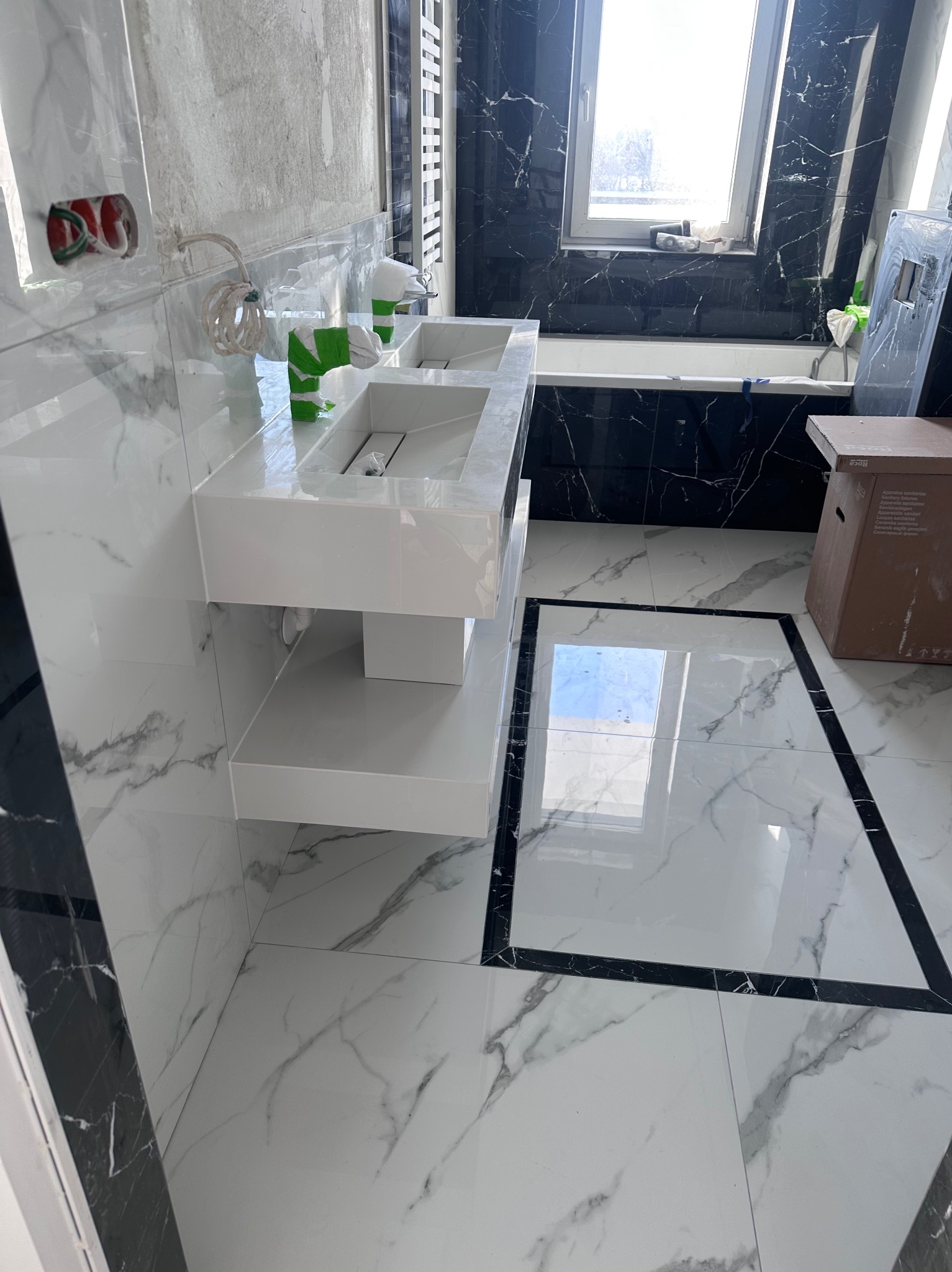 Nowoczesna łazienka z marmurowymi płytkami i białą, podwieszaną umywalką. Widoczne wykończenia w czarnym marmurze i fragment okna.