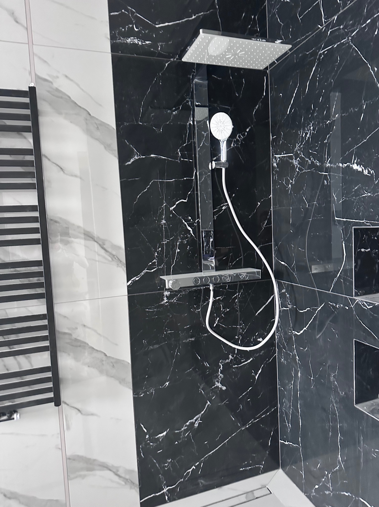 Nowoczesna kabina prysznicowa z czarnymi płytkami imitującymi marmur i deszczownicą. Widoczny grzejnik drabinkowy i białe płytki z szarymi żyłami.