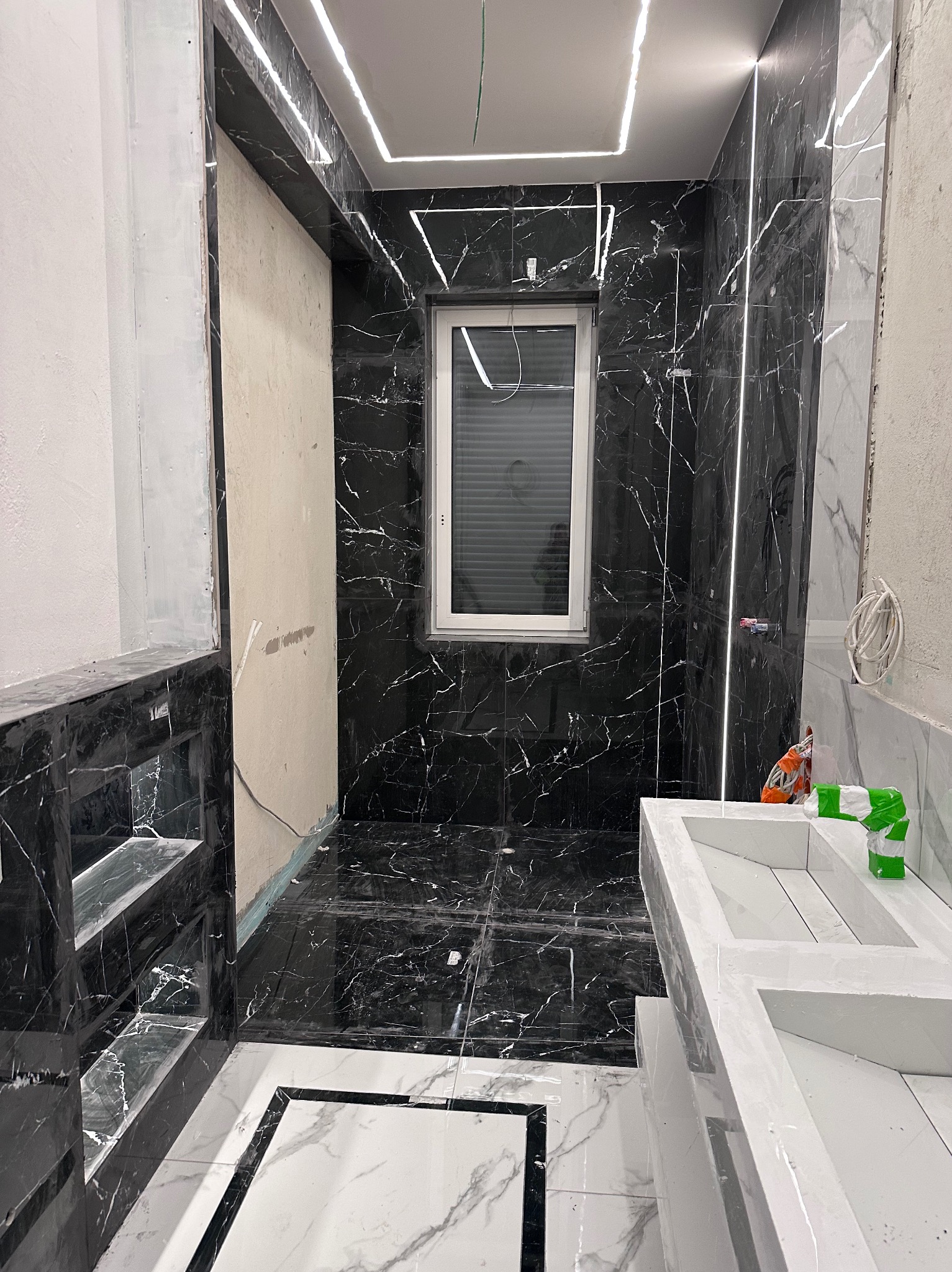 Częściowo wykończona łazienka z czarnymi płytkami imitującymi marmur, białymi akcentami i oświetleniem LED. Widoczna umywalka i półki wnękowe.