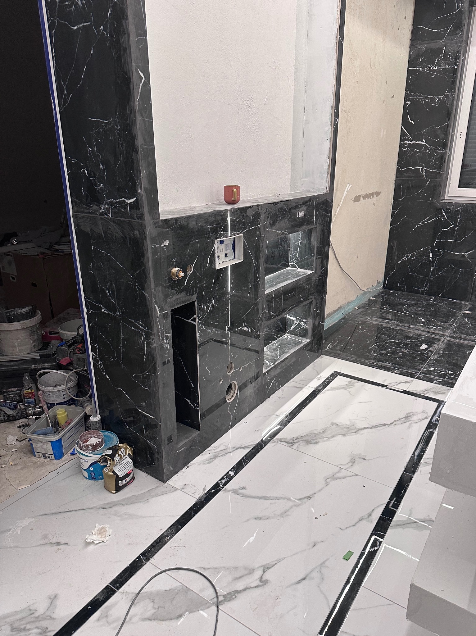 Łazienka w trakcie wykańczania: czarne płytki imitujące marmur na ścianach i zabudowie, białe płytki podłogowe z czarnymi akcentami. Widoczne narzędzia i materiały budowlane.