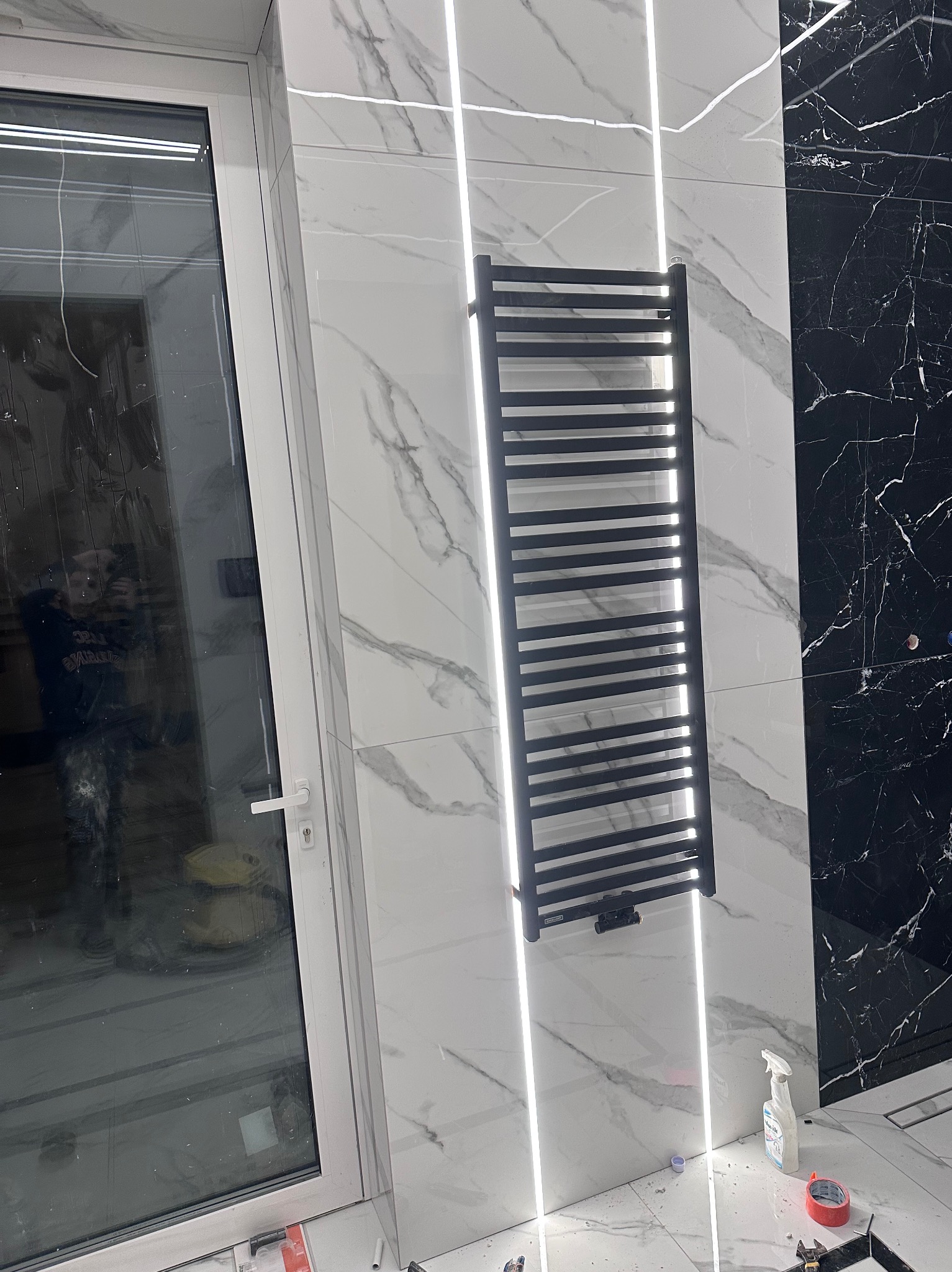 Nowoczesna łazienka z czarno-białymi marmurowymi płytkami, podświetleniem LED i czarnym grzejnikiem drabinkowym. Widoczne akcesoria montażowe.