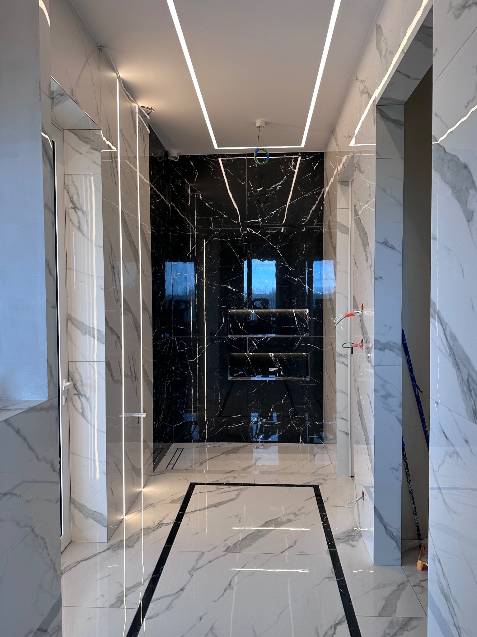 Nowoczesna łazienka z czarno-białymi marmurowymi płytkami, podświetleniem LED i szklaną kabiną prysznicową. Widoczne elementy instalacji elektrycznej.