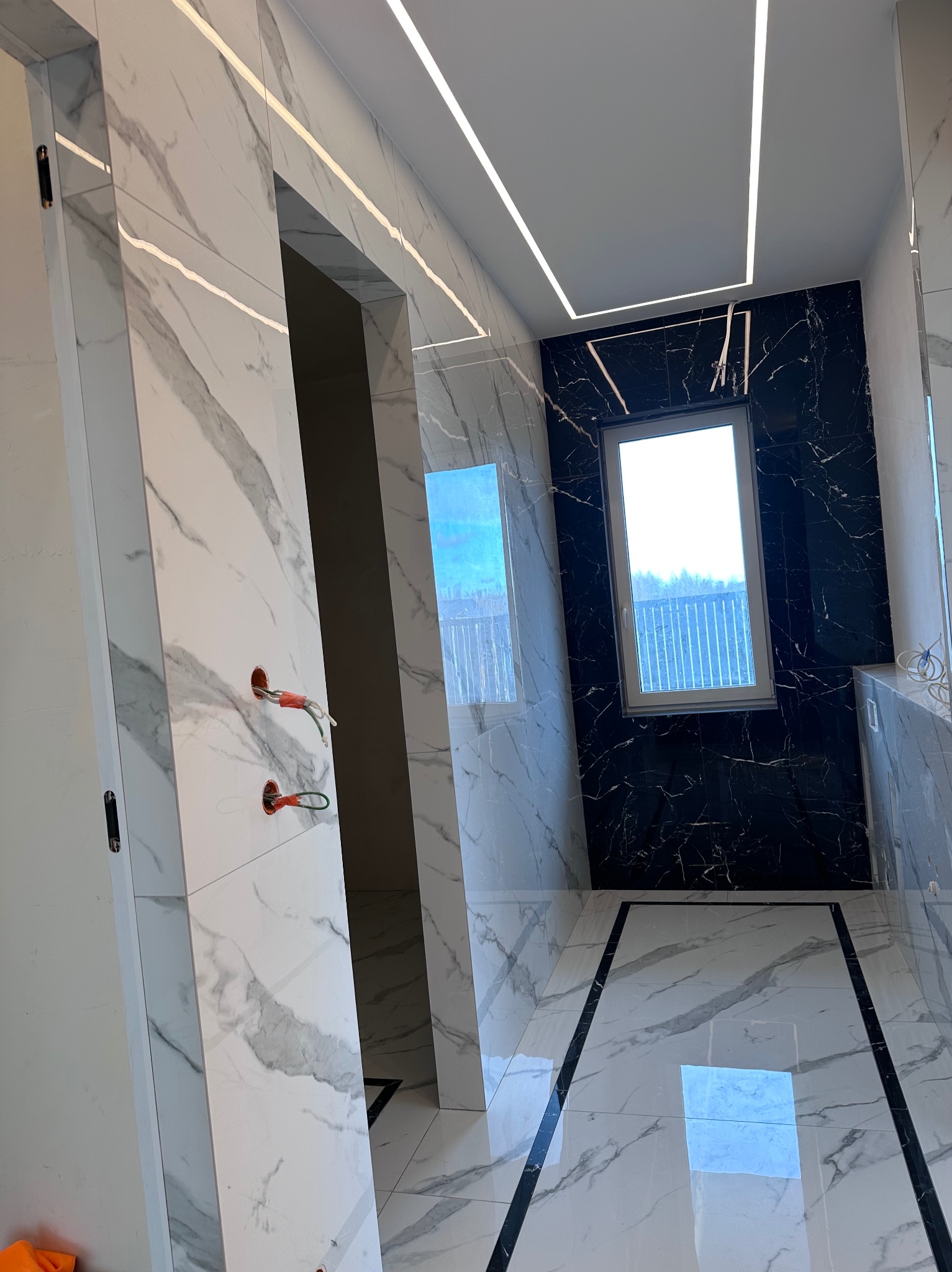 Nowoczesna łazienka z marmurowymi płytkami w odcieniach bieli i czerni, podświetlanym sufitem i oknem. Widoczne niedokończone instalacje elektryczne.