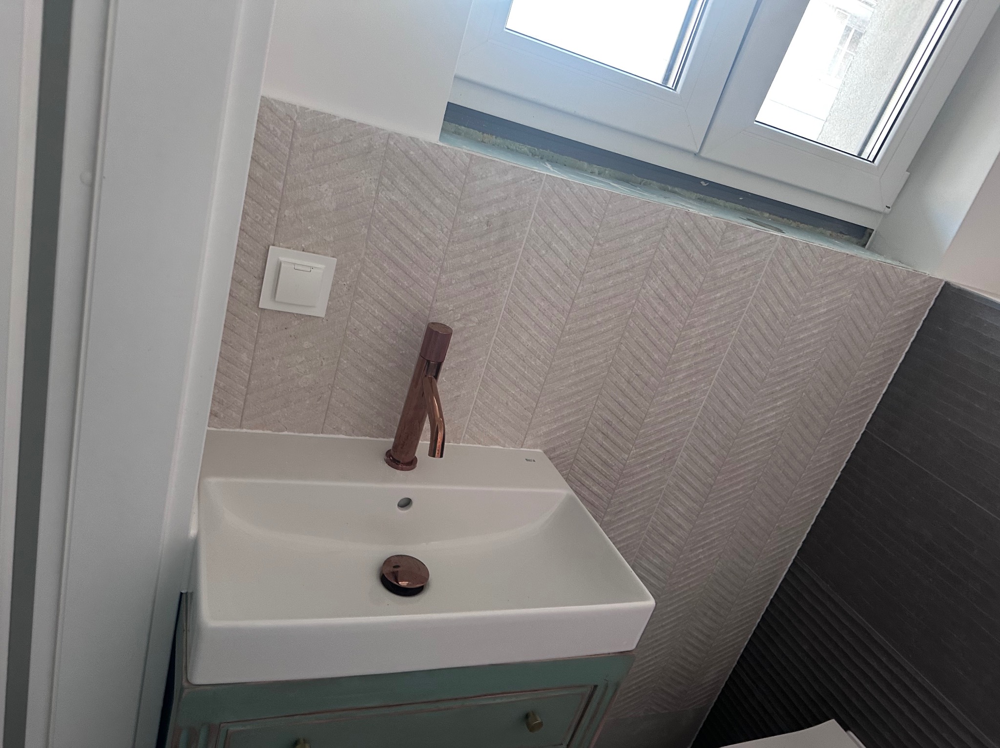 Umywalka z szafką w stylu retro, bateria w kolorze miedzi, płytki w jodełkę na ścianie, okno w tle. Wykończona łazienka z dbałością o detale.