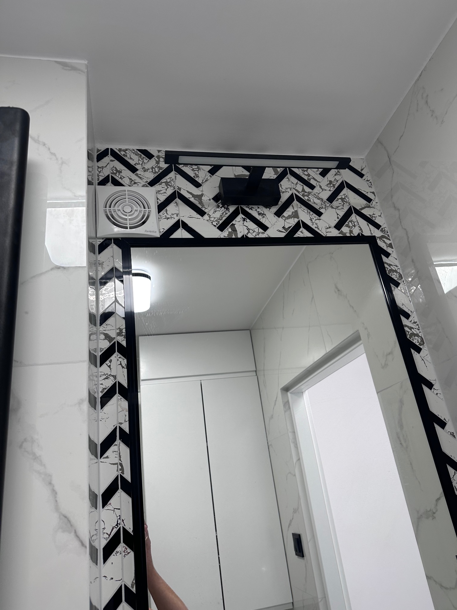 Nowoczesna łazienka z lustrem w czarnej ramie, otoczonymi płytkami w zygzak. Widoczny wentylator i oświetlenie nad lustrem. Białe marmurowe płytki na ścianach.