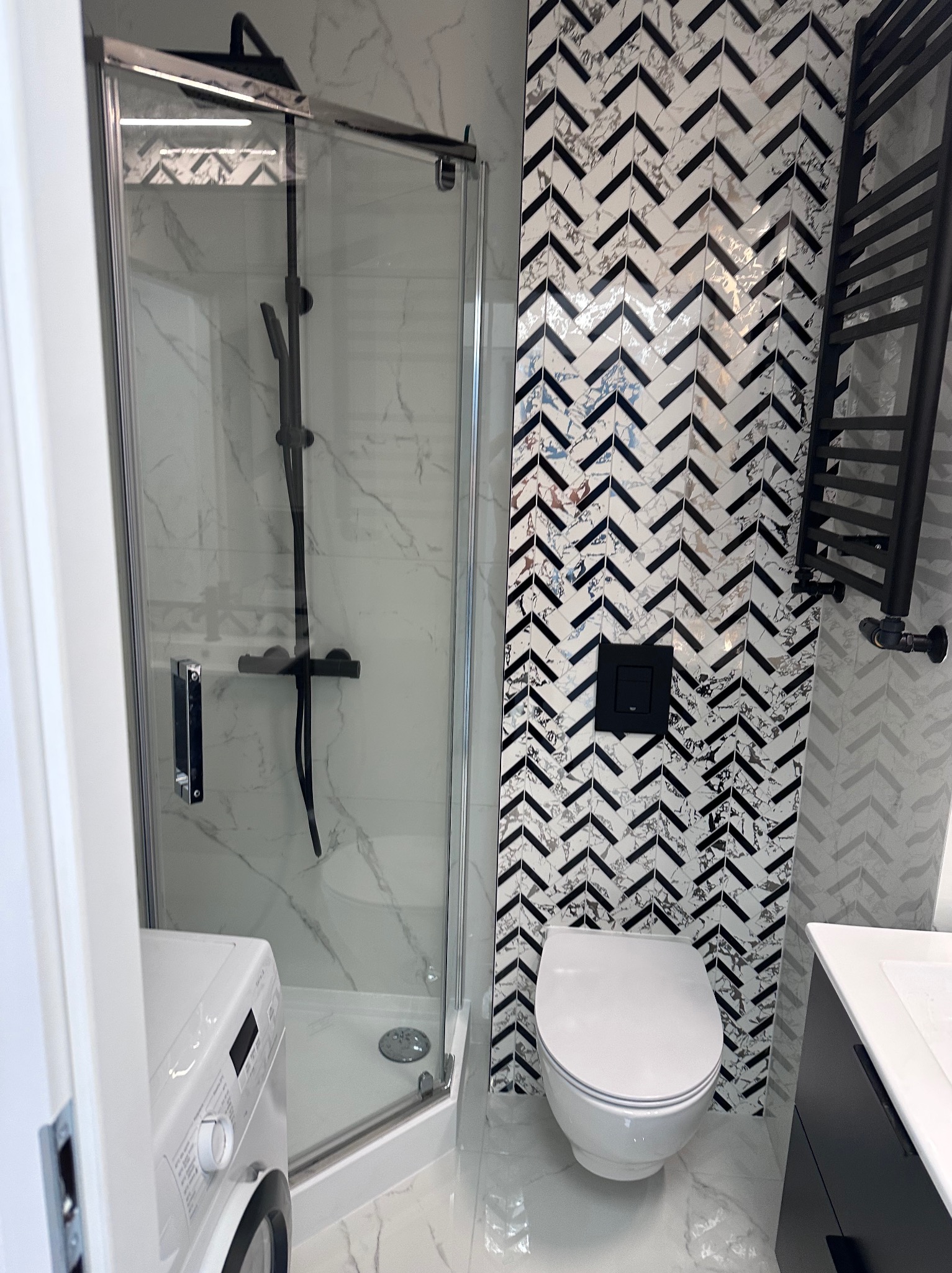 Nowoczesna łazienka z kabiną prysznicową typu walk-in, toaletą podwieszaną i grzejnikiem drabinkowym w czarnym kolorze. Uwagę przyciągają płytki ułożone w jodełkę.