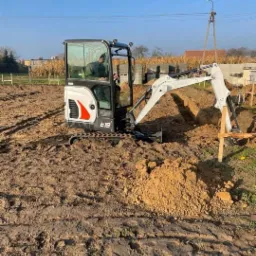 Mini koparka Bobcat E19 w trakcie pracy na placu budowy, tworząca wąski wykop w ziemi, z widocznymi śladami gąsienic i stertą wykopanej ziemi obok, w tle widoczne bloczki fundamentowe i słup...