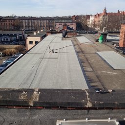 PAMADACH Paweł Maciejczyk - Praca na płaskim dachu: dekarz w trakcie układania papy termozgrzewalnej, widoczny palnik i butla gazowa. W tle zabudowa miejska i błękitne niebo.