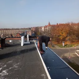 Praca na dachu płaskim: dekarz mocuje papę termozgrzewalną wkrętarką. W tle zabudowa miejska i jesienne drzewa. Widok z góry na etap krycia dachu.