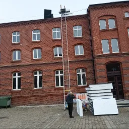 Pracownik na dachu ceglanego budynku, używa podnośnika i montuje panele. Stos paneli leży obok drabiny. Widoczny fragment elewacji i brukowana nawierzchnia.