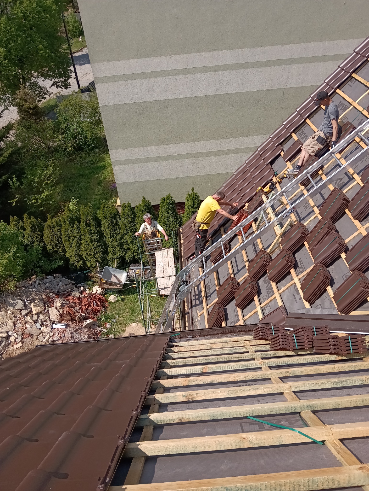 Montaż brązowej dachówki na drewnianej konstrukcji dachu. Widoczni dekarze na drabinie i rusztowaniu. Fragment budynku w tle. Ujęcie z góry.
