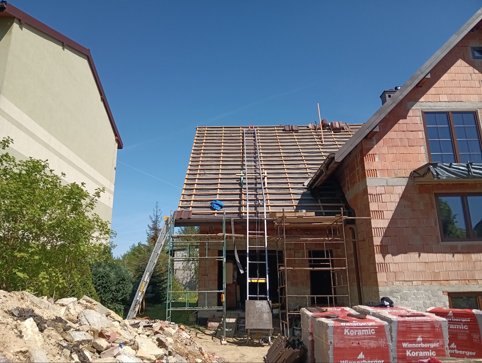 Budowa dachu z dachówki ceramicznej Wienerberger na domu jednorodzinnym. Widoczne rusztowania i drabiny. Częściowo ułożona dachówka na łatach.