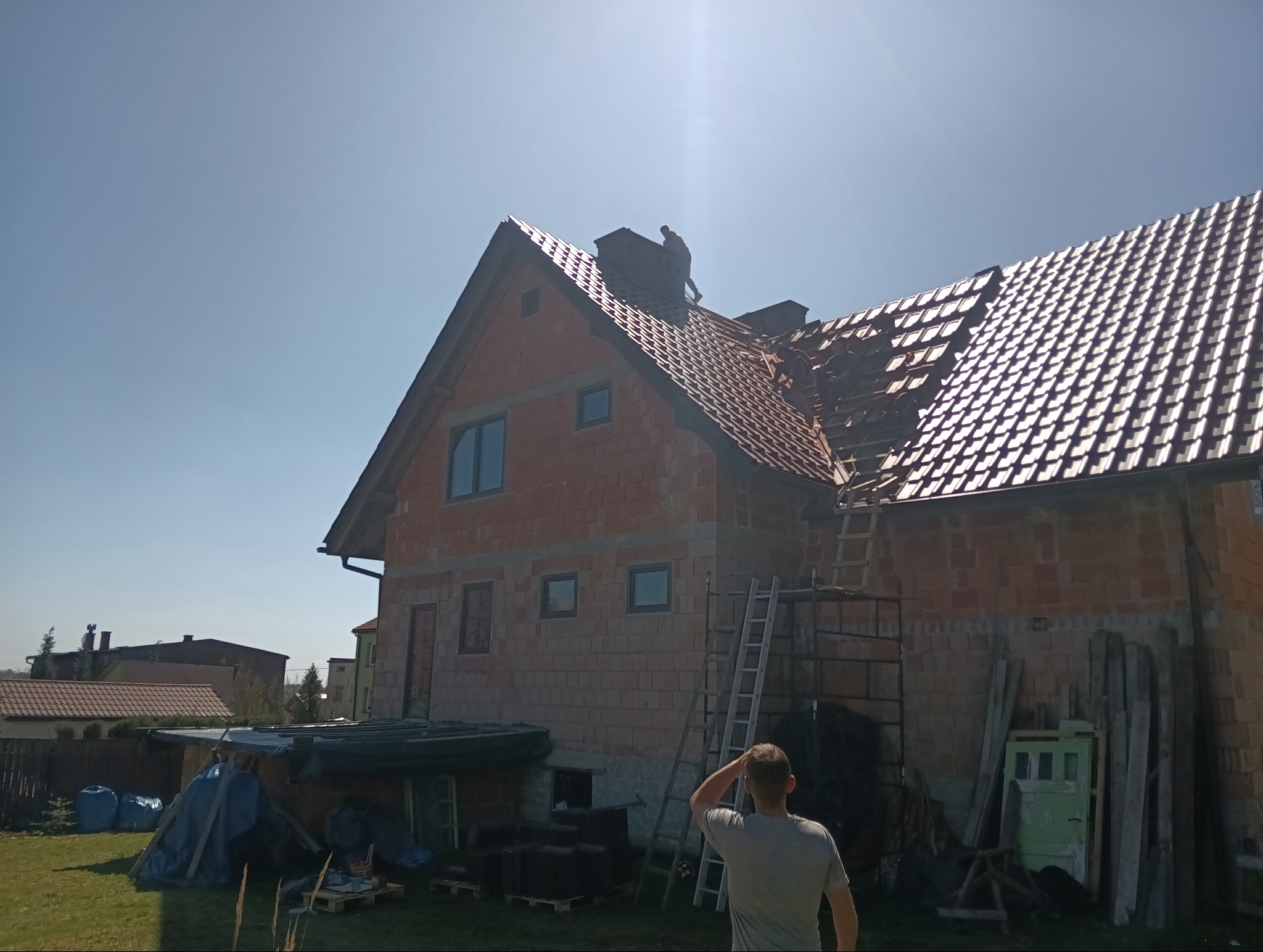 Ceglany dom w trakcie krycia dachu czerwoną dachówką. Widoczny dekarz na szczycie dachu i drabina oparta o ścianę. Częściowo pokryty dach. Słoneczny dzień.