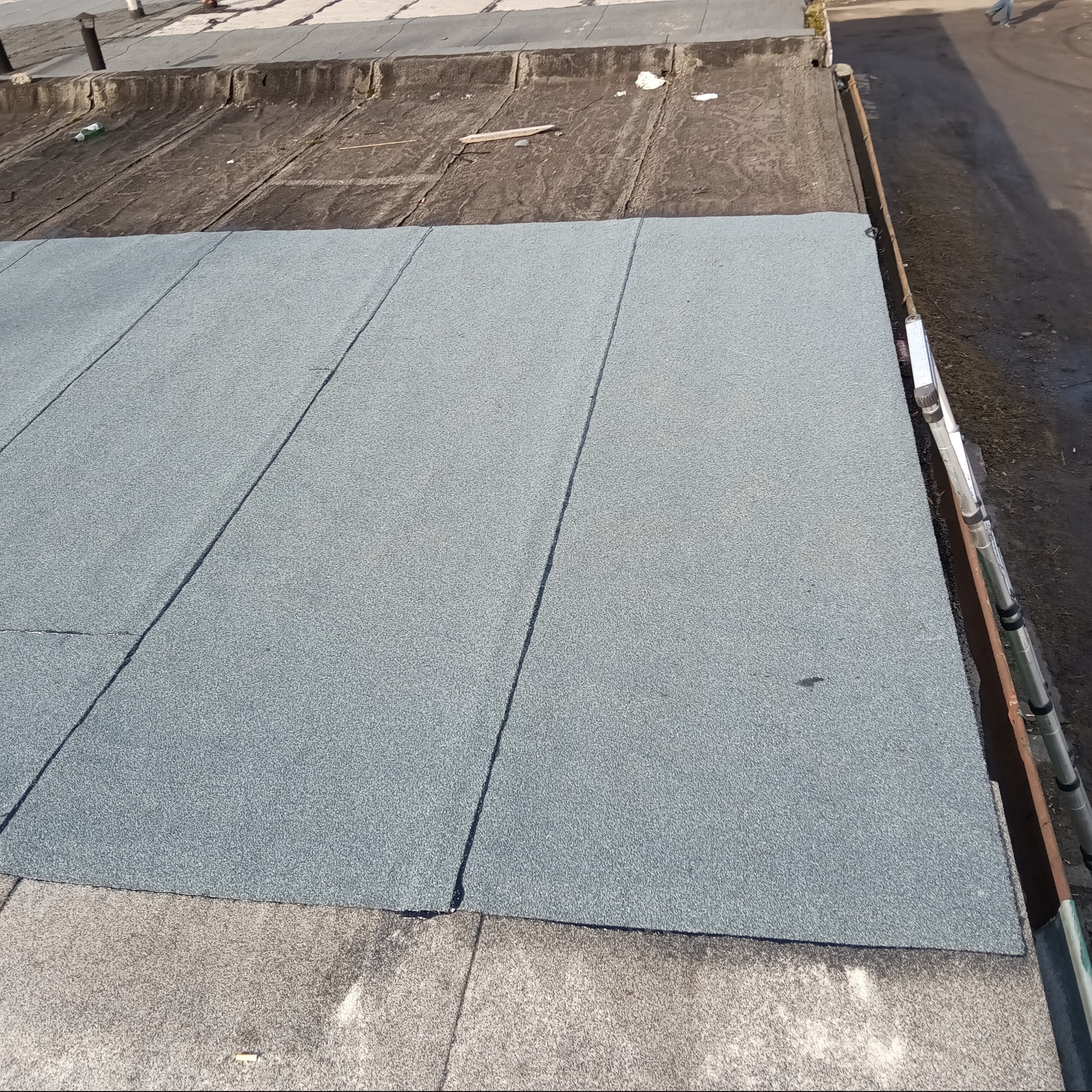 Fragment płaskiego dachu pokrytego nową papą w kolorze szarym, z widocznymi łączeniami. W tle stara, uszkodzona powierzchnia dachu i drabina oparta o krawędź.