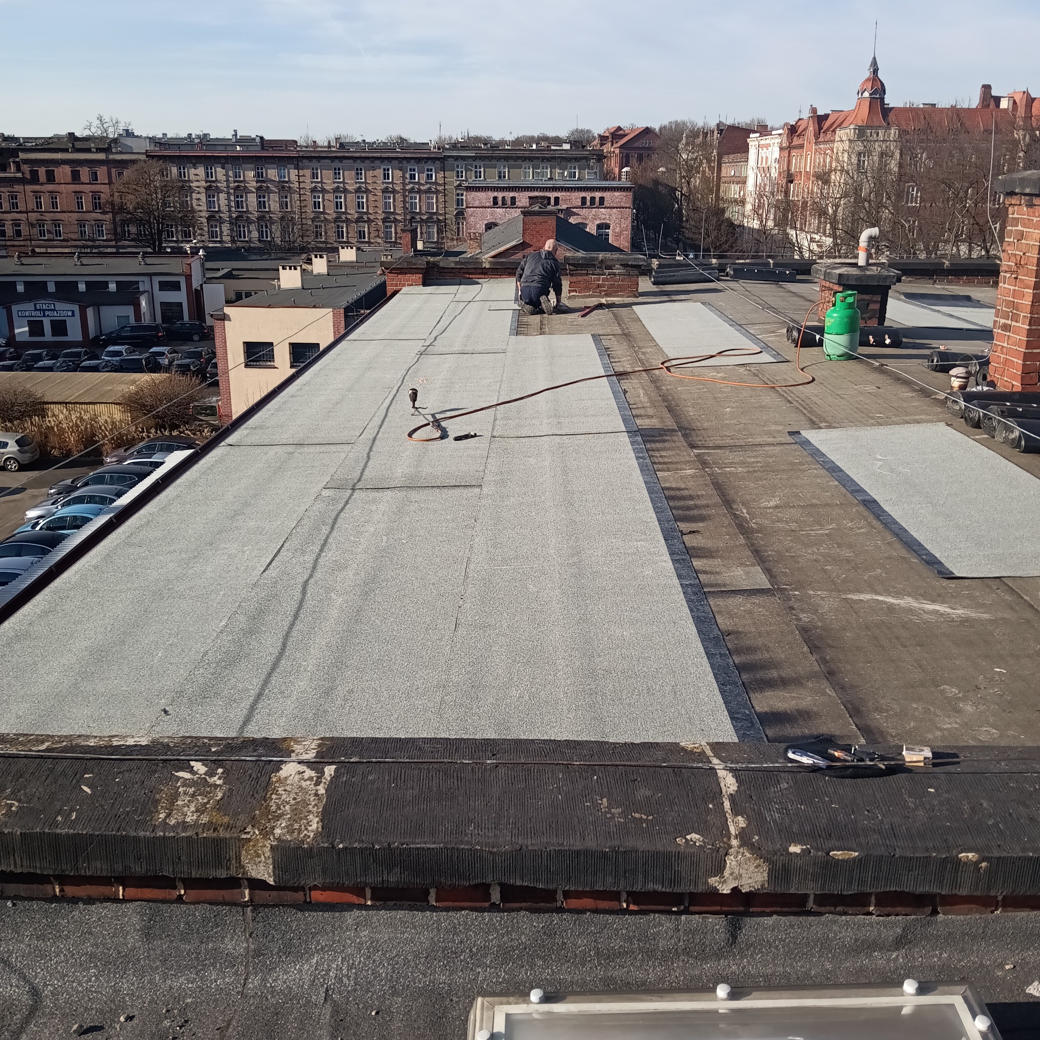 Praca na płaskim dachu: dekarz w trakcie układania papy termozgrzewalnej, widoczny palnik i butla gazowa. W tle zabudowa miejska i błękitne niebo.