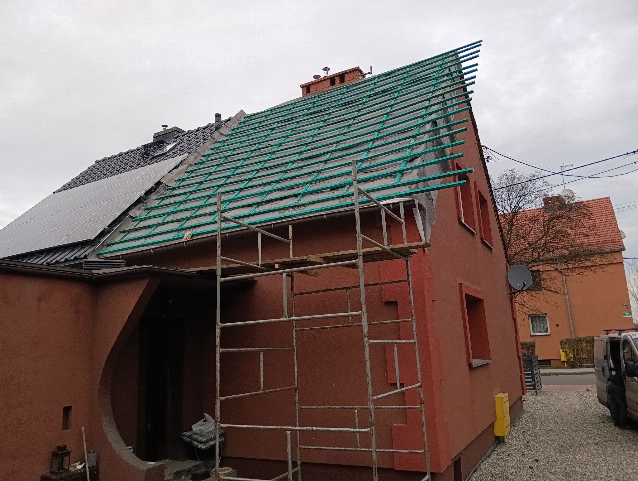Częściowo zdjęty dach z łatami i folią w kolorze zielonym, widoczne rusztowanie i panele fotowoltaiczne na budynku w trakcie remontu.