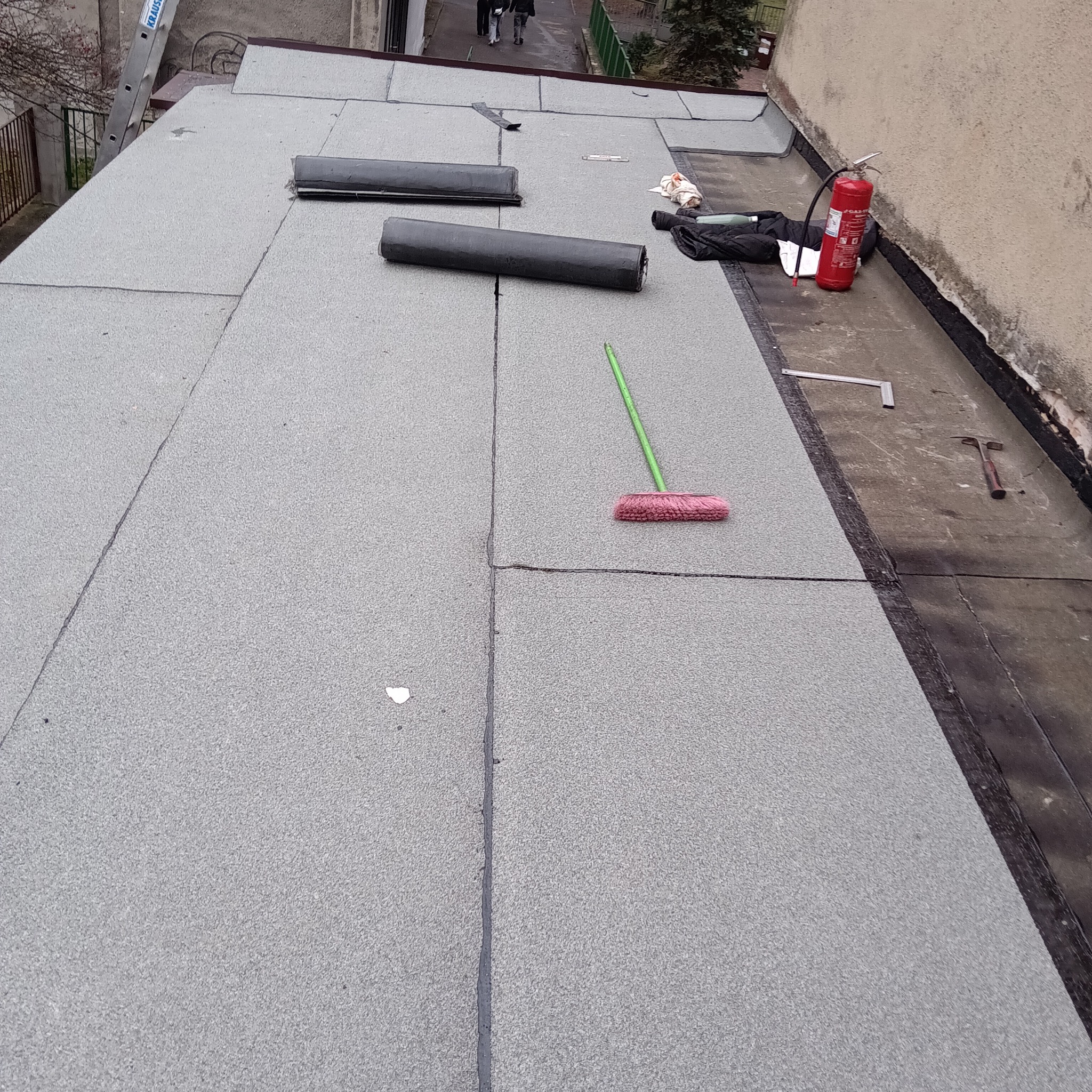 Fragment dachu pokrytego papą termozgrzewalną. Widoczne narzędzia: szczotka, młotek, kątownik. Na dachu leżą rolki papy i gaśnica.