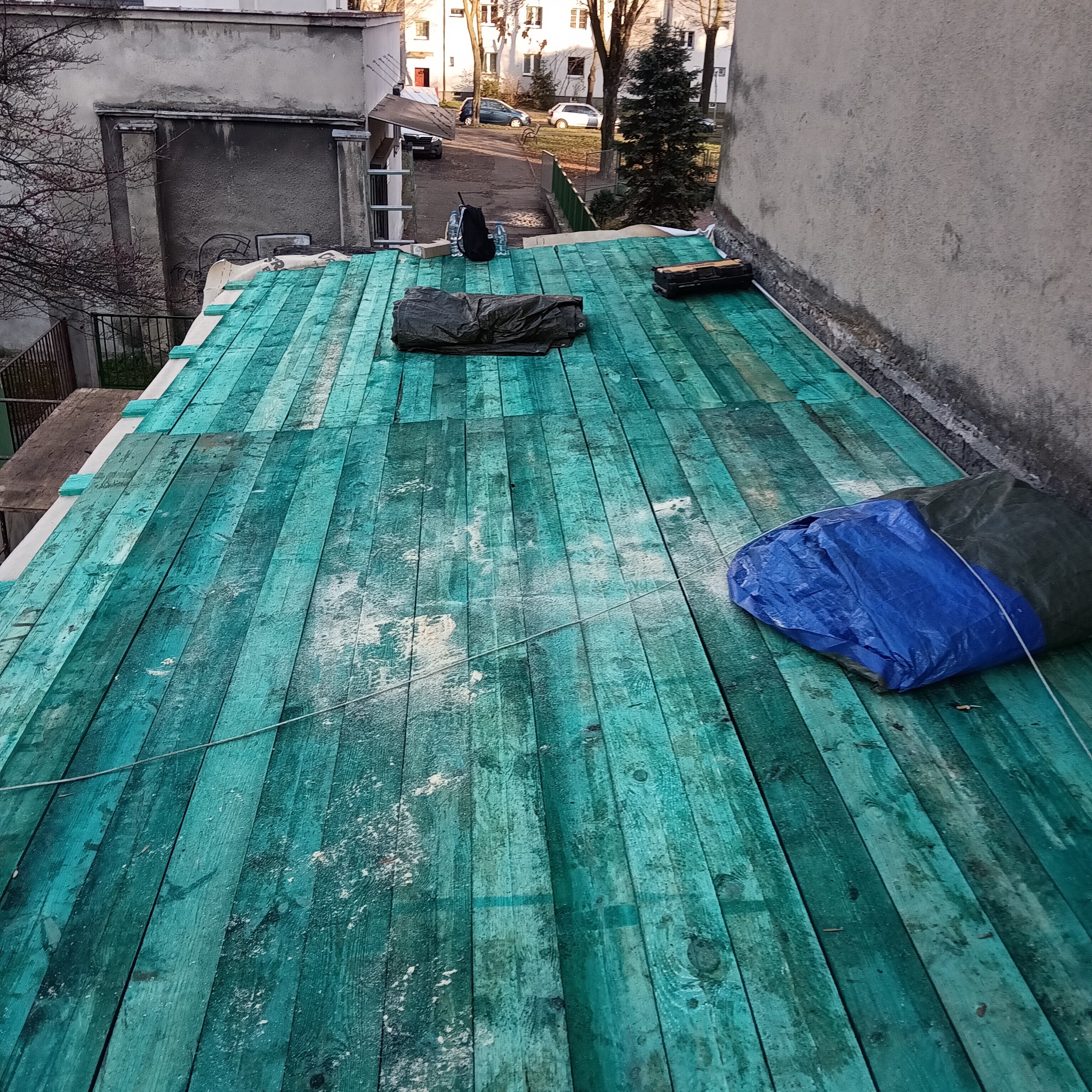 Zielone deski na dachu budynku w trakcie prac dekarskich. Widoczne narzędzia i materiały budowlane. Perspektywa z góry ukazuje strukturę dachu i otoczenie.