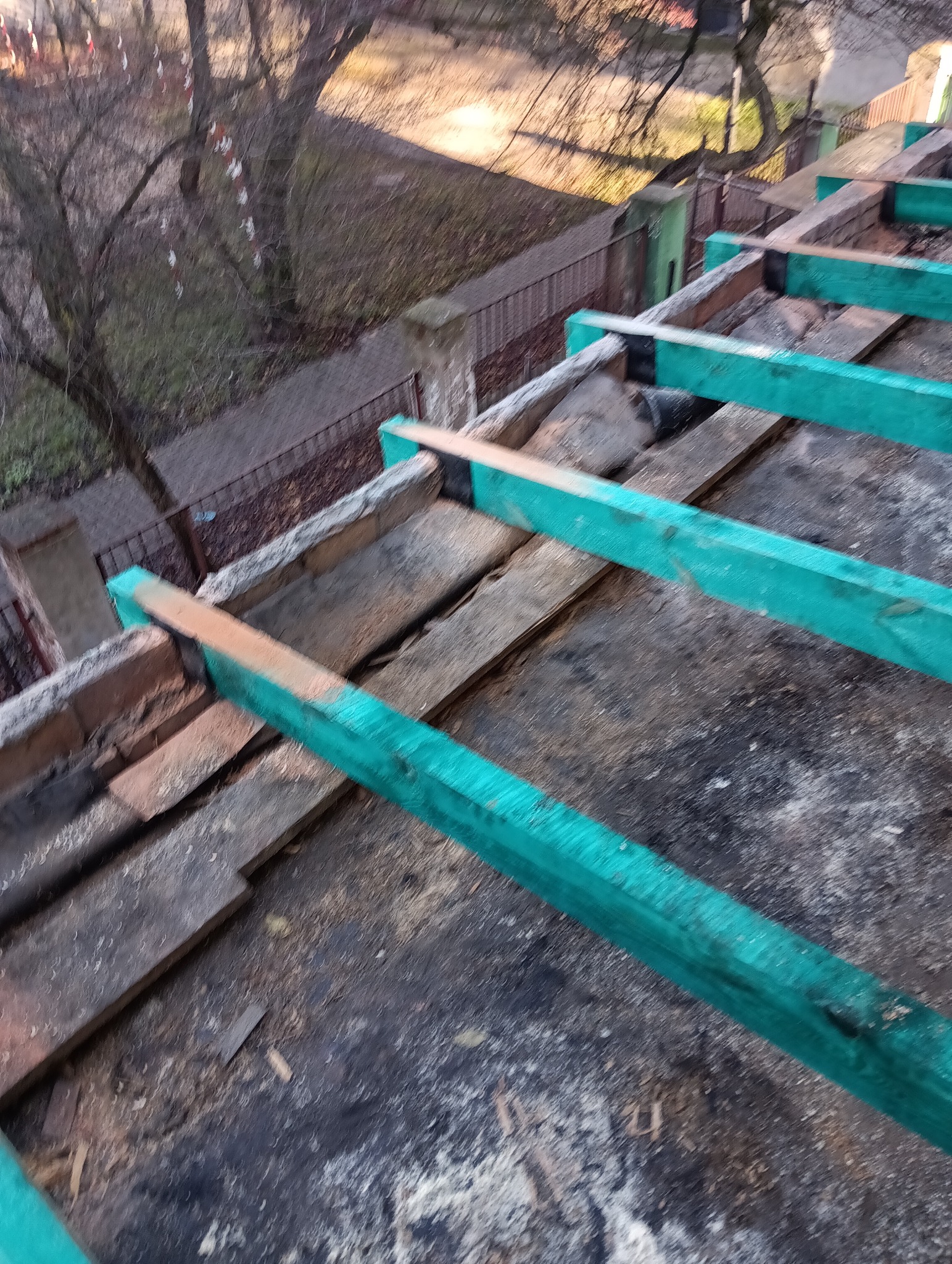 Demontaż dachu z widocznymi elementami konstrukcyjnymi i zielonymi belkami, z perspektywy góry, w tle widoczny budynek i drzewa.