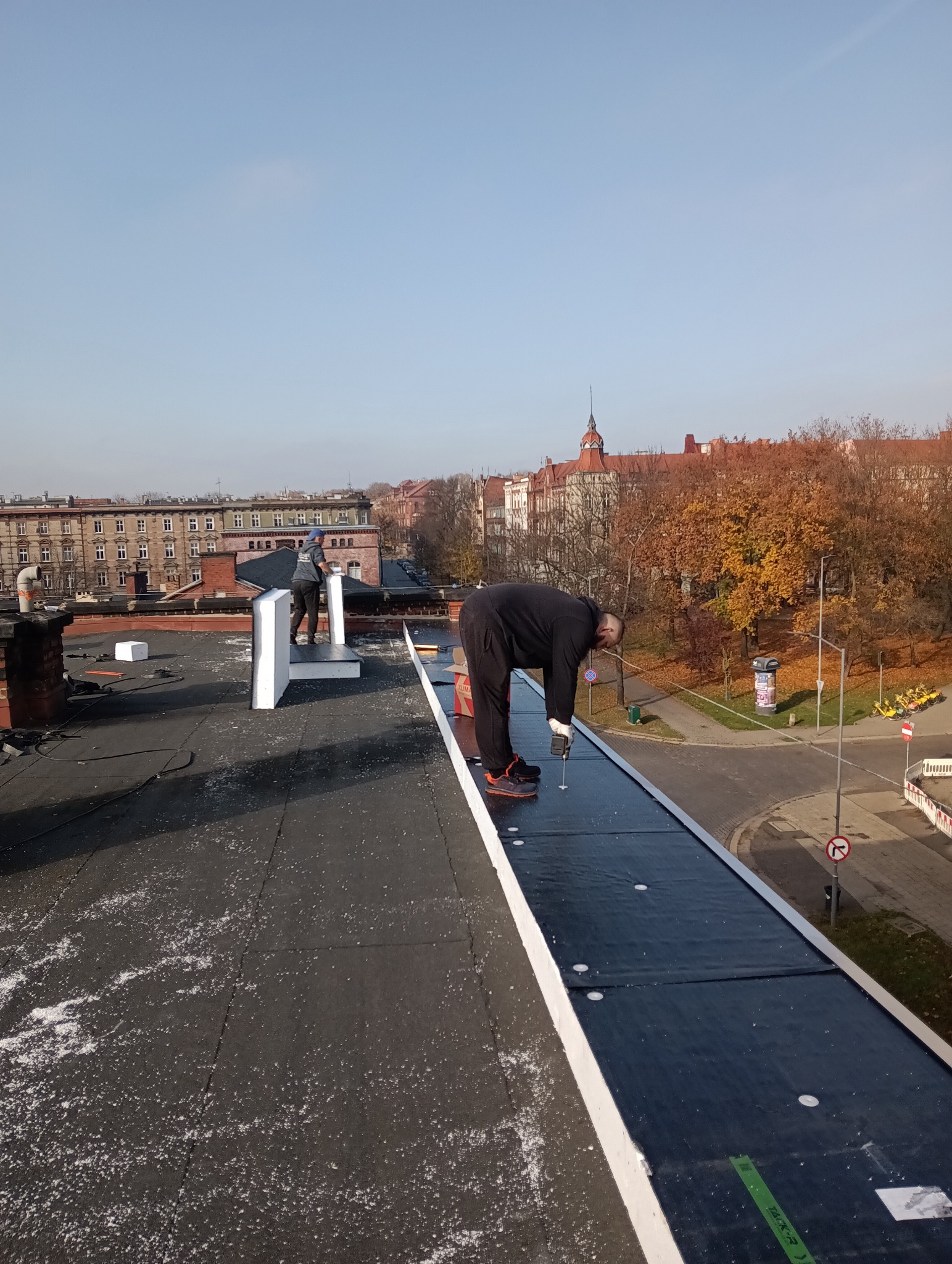 Praca na dachu płaskim: dekarz mocuje papę termozgrzewalną wkrętarką. W tle zabudowa miejska i jesienne drzewa. Widok z góry na etap krycia dachu.