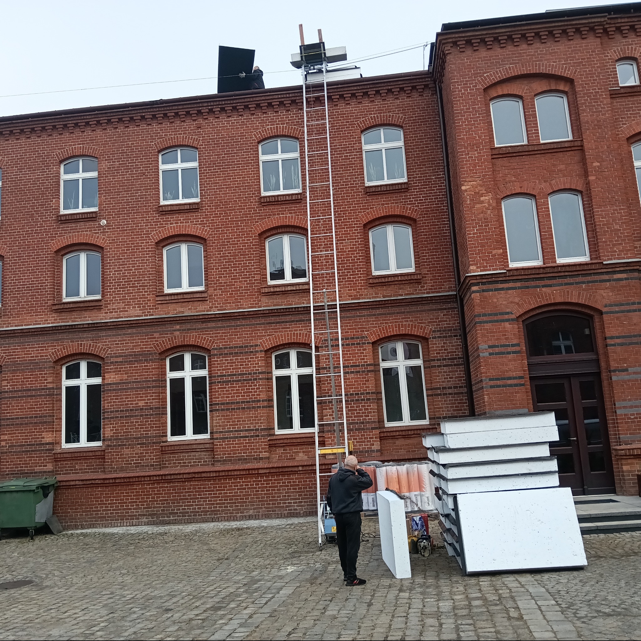 Pracownik na dachu ceglanego budynku, używa podnośnika i montuje panele. Stos paneli leży obok drabiny. Widoczny fragment elewacji i brukowana nawierzchnia.