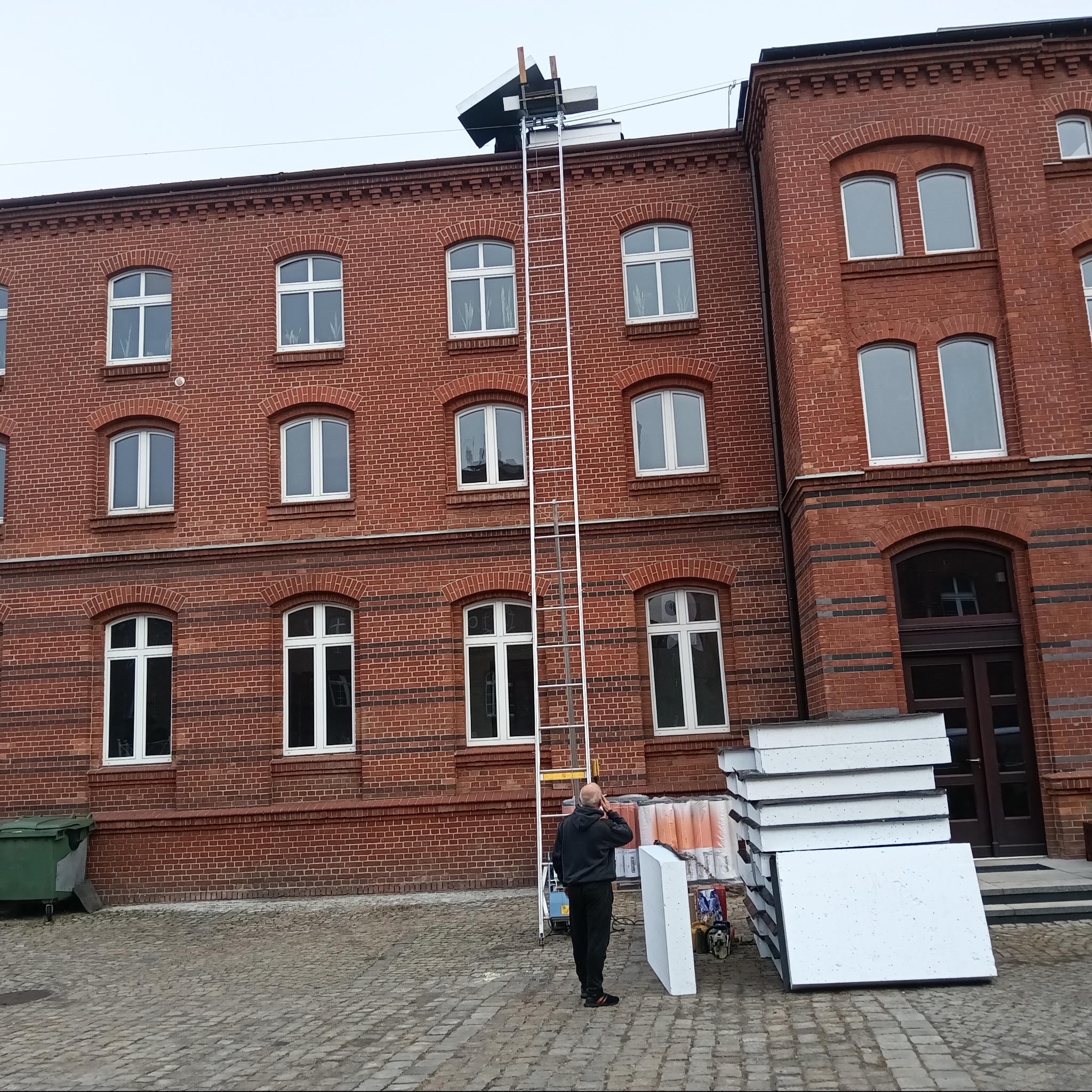 Prace na dachu ceglanego budynku. Osoba rozmawia przez telefon obok styropianu i drabiny. Na dachu widoczne elementy konstrukcji. Prace dekarskie w toku.