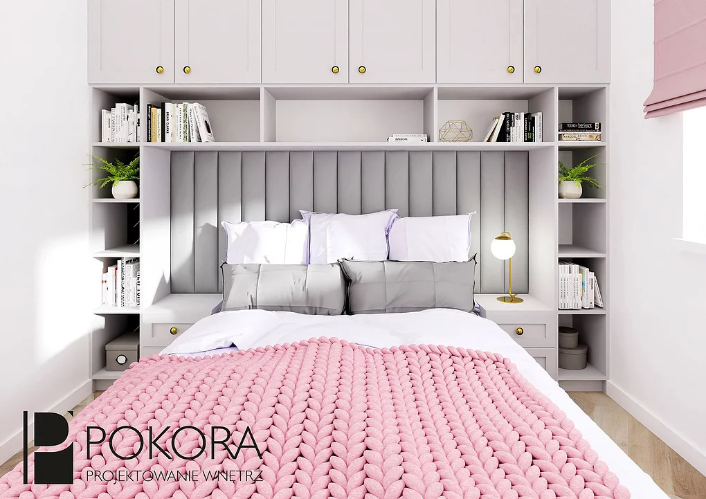 Jasna sypialnia z zabudową meblową nad wezgłowiem łóżka, szara tapicerowana ściana, różowy pled z grubej wełny i złota lampka nocna.