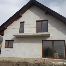 Surowa elewacja domu jednorodzinnego z brązowymi oknami, widoczne zarysowania na ścianie, niezakończone prace wykończeniowe wokół budynku.