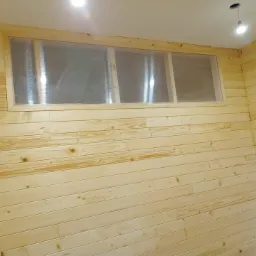 Wnętrze wykończone jasnymi panelami drewnianymi ułożonymi poziomo, z oknem z przezroczystą, żebrowaną szybą i widoczną konstrukcją ramy, oraz punktowym oświetleniem sufitowym.