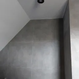 Ściana w łazience wyłożona szarymi płytkami w formacie kwadratu, widoczny fragment sufitu ze skośną częścią i okrągłym oświetleniem oraz fragment drewnianej ramy drzwi.