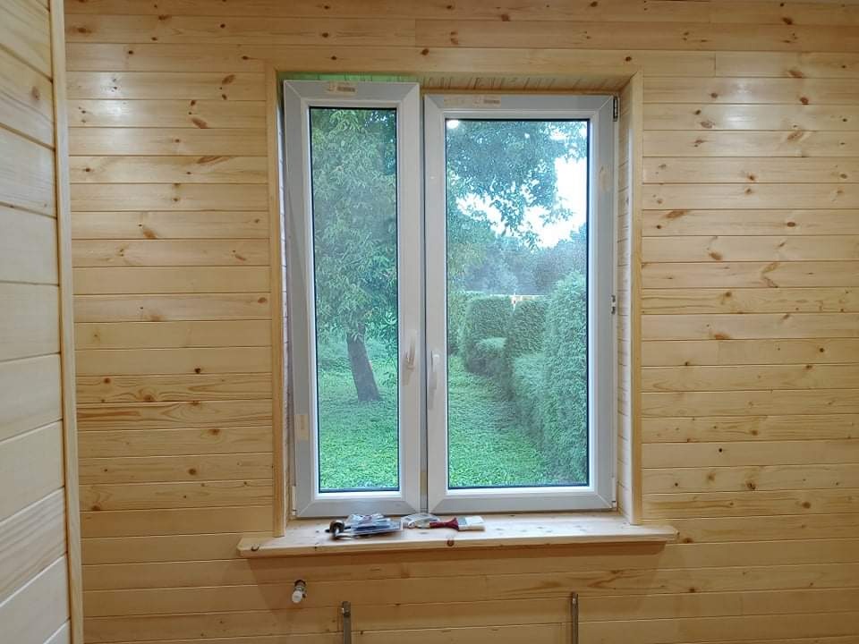 Wnętrze wykończone boazerią drewnianą z białym oknem PCV, widok na zielony ogród z formowanymi krzewami, narzędzia na parapecie.