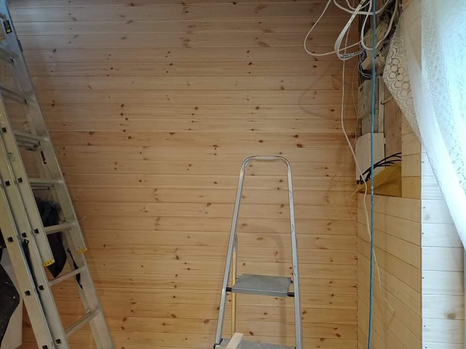 Ściana wyłożona boazerią z widoczną instalacją elektryczną i dwoma różnymi drabinami, jedna oparta o ścianę, druga stojąca na środku.