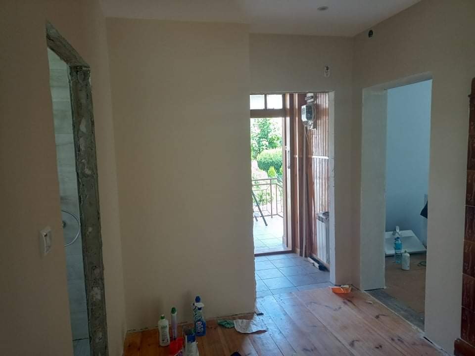 Wnętrze po świeżym malowaniu ścian na jasny beż, z widocznymi otworami drzwiowymi, drewnianą podłogą i butelkami z detergentami w rogu pokoju.