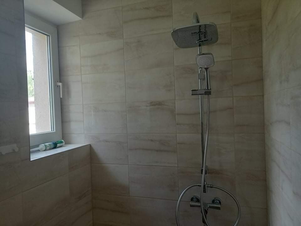 Wykończona łazienka z beżowymi płytkami imitującymi kamień, nowoczesna kabina prysznicowa z deszczownicą i widocznym silikonem sanitarnym przy oknie.