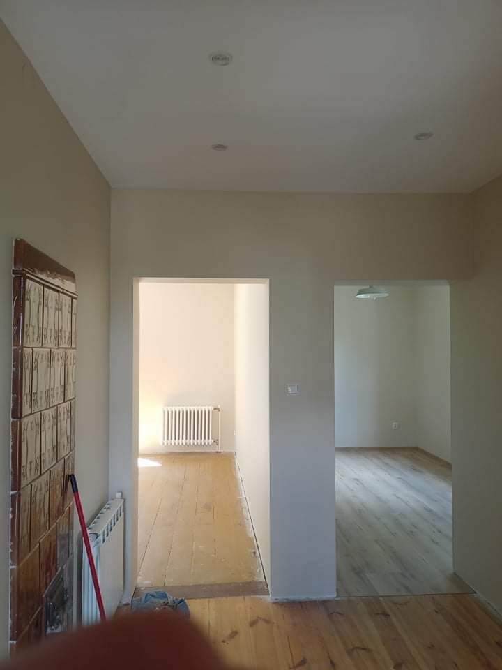 Wnętrze mieszkania w trakcie odświeżania: pomalowane ściany w odcieniach beżu, widoczne dwa przejścia do innych pomieszczeń z grzejnikami. Podłoga częściowo odnowiona, z widocznymi śladami starej...