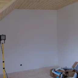 Pokój na poddaszu w trakcie prac wykończeniowych: świeżo pomalowane ściany na biało, drewniana boazeria na skosach, kartony po płytkach na podłodze i lampa budowlana na statywie.