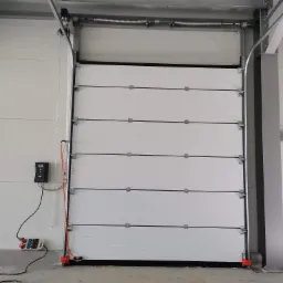 Zamknięta, biała segmentowa brama przemysłowa z widocznym mechanizmem sprężynowym, łańcuchem, sterownikiem i zabezpieczeniami na dole, zamontowana w hali.