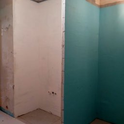 Wiesław Hojdak - Dwie ściany łazienki w trakcie remontu: jedna w surowym stanie z widocznymi instalacjami, druga pomalowana na turkusowo, z nierównym paskiem surowego tynku u góry. Widoczna podłoga z płytek...