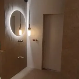 Nowoczesna łazienka w beżowych odcieniach z okrągłym lustrem podświetlanym LED, minimalistyczną armaturą i wiszącymi lampami kulistymi.