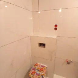 Wykończona łazienka z białymi, marmurkowymi płytkami na ścianach i podłodze, z toaletą z zabudowaną spłuczką i deską klozetową z motywem komiksowym.