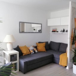 Wiesław Hojdak - Jasne wnętrze pokoju dziennego z szarą narożną sofą, żółtymi poduszkami i kocem, białym stolikiem z lampką, lustrem w szarej ramie i roślinami w doniczkach, całość na jasnej podłodze.
