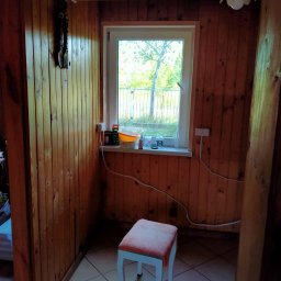 Wiesław Hojdak - Wnętrze pokoju wykończonego boazerią, z oknem, stołkiem z pomarańczowym siedziskiem i ceramiczną podłogą w kratkę. Na parapecie okna widoczne pojemniki i puszki.