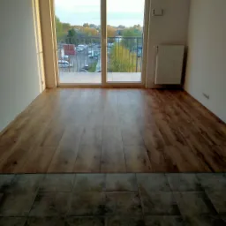 Fragment pokoju z nową podłogą: panele imitujące drewno w jasnym odcieniu przechodzące w ciemne, kwadratowe płytki. Widok na balkon z metalową barierką i parkingiem w tle, przez przesuwne drzwi...