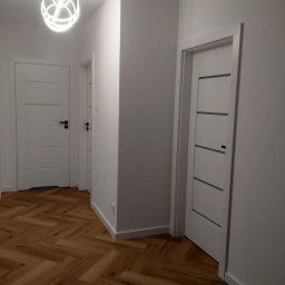 Wiesław Hojdak - Korytarz po remoncie: Świeżo pomalowane białe ściany, dwie białe drzwi z czarnymi klamkami i szarymi wstawkami, parkiet ułożony w jodełkę francuską, nowoczesna lampa sufitowa.